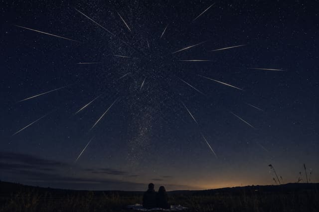 The Perseid meteor shower over a dark horizon, trails radiating from a single point, two people watching from a blanket.